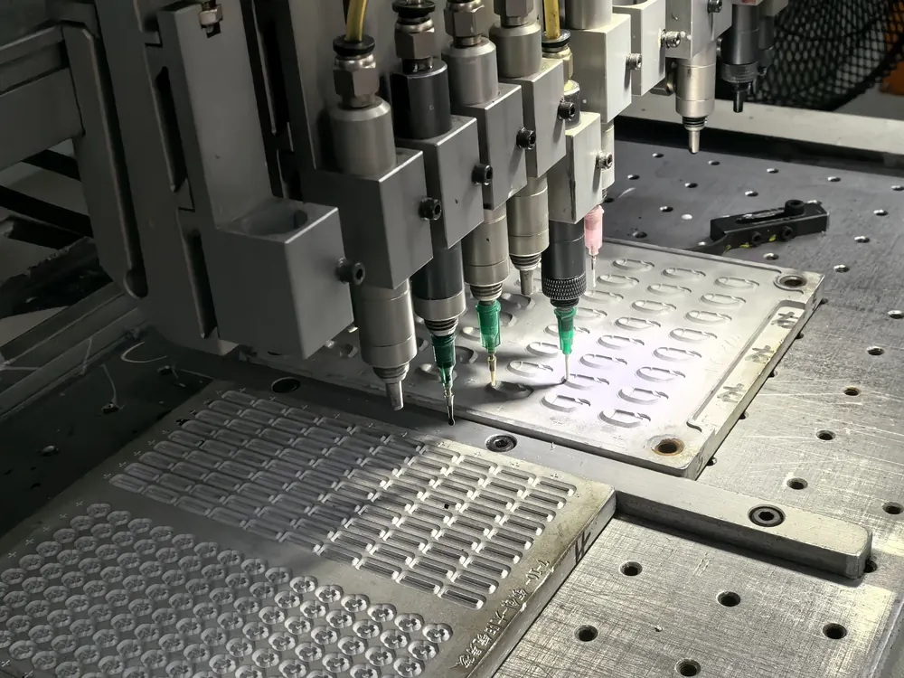 Multi-nozzle dispensing head laying beads of coloured silicone into an aluminium mould cavity.